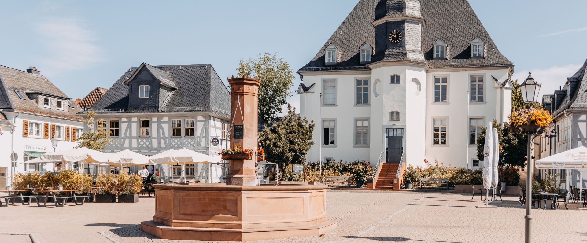 Alter Marktplatz in Usingen Alter Marktplatz in Usingen mit großem Sandsteinbrunnen und historischem Gebäude mit Uhrturm. Fachwerkhäuser, Außengastronomie, Blumen und Sitzbänke umgeben den gepflasterten Platz.