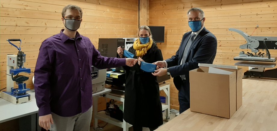Links steht Herr Ralf Nikel von Youkon, welcher mit Bürgermeister Herr Steffen Wernard eine der neuen blauen Usinger Masken hochhält. Leicht nach hinten versetzt steht Personalabteilungsleiterin Frau Vanessa Knull, welche ebenfalls eine blaue Usinger Maske hochhält. 