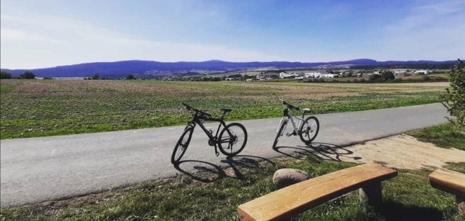 Im Hintergrund befindet sich eine idyllische Landschaft mit grüner Wiese. Im Vordergrund stehen zwei Fahrräder und eine Sitzbank aus Holz.
