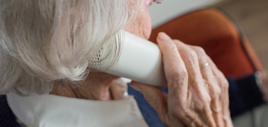 Eine ältere Dame, welche von der seite abgelichtet wurde. Sie hält einen weißen Telefonhörer mit ihrer rechten Hand an ihr rechtes Ohr. 