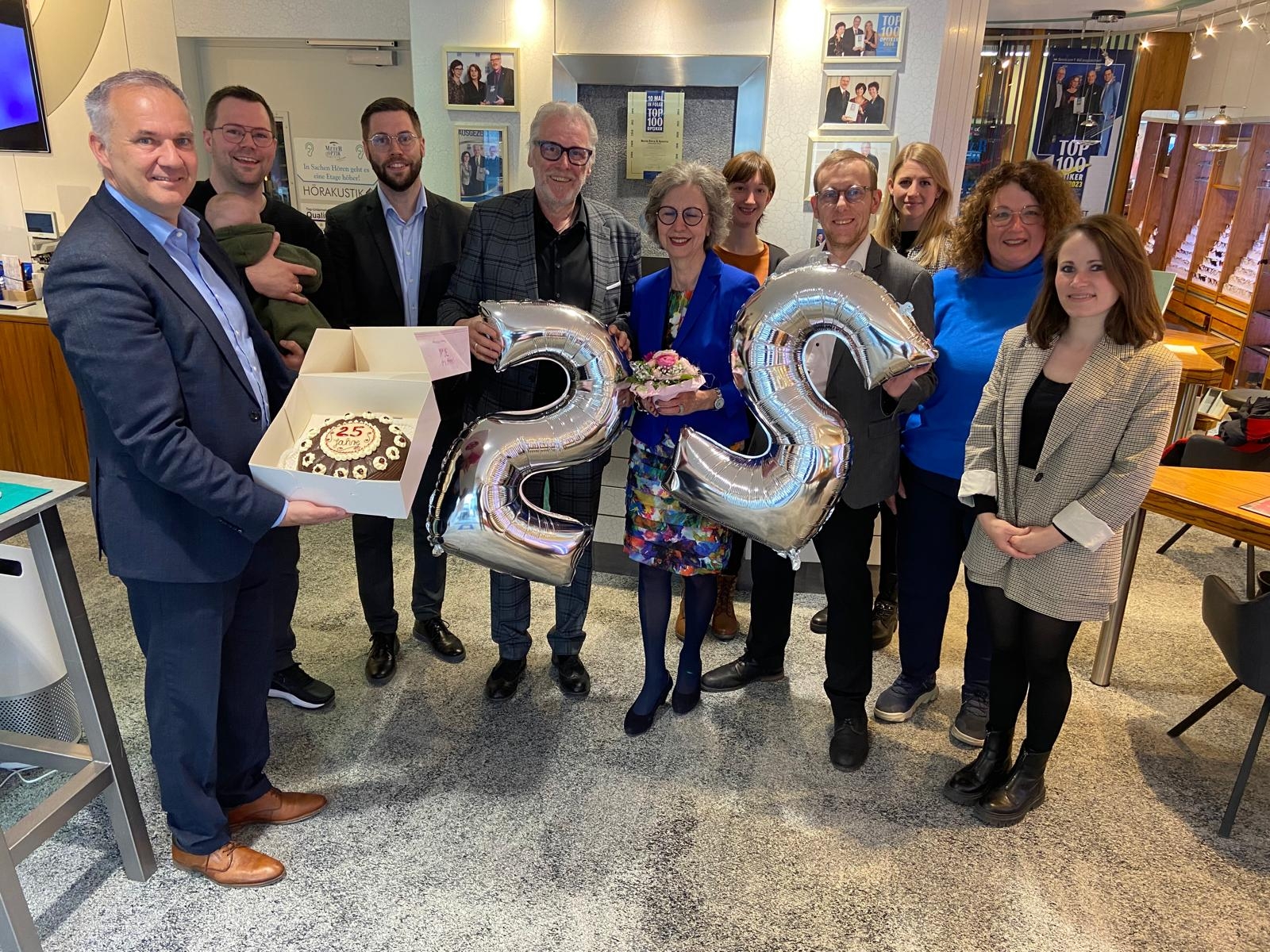 Das Bild zeigt eine Gruppe von elf Personen, die gemeinsam ein Jubiläum feiern. Zwei Personen halten silberne Ballons in Form der Zahl 25, während ein Mann eine Torte in einem Karton präsentiert. Zum Jubiläum gratulierten Bürgermeister Steffen Wernard und Wirtschaftsförderin Ute Harmel mit einer leckeren Haustorte vom benachbarten Schlosscafé Keth und bedankten sich zudem für das verlässliche Engagement von Familie Jenisch und dem Team von Meyer Optik & Akustik bei Innenstadt- und Werbeaktivitäten sowie der verlässlichen Unterstützung von Charity- und Vereinsveranstaltung in Usingen.