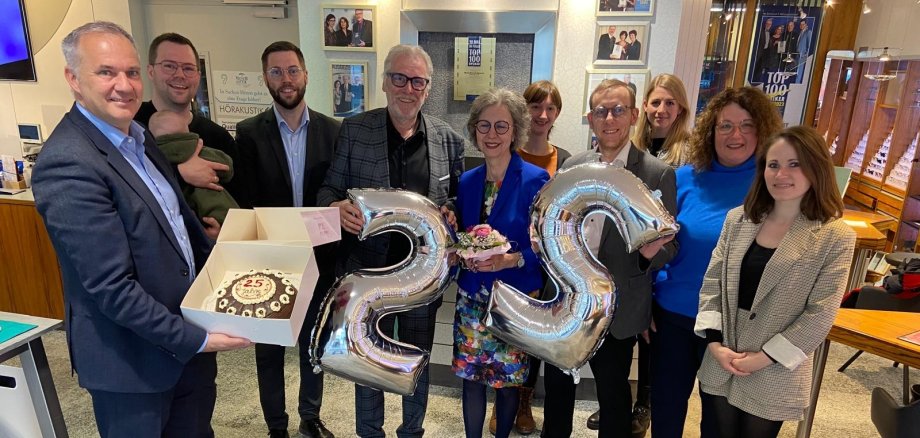 Das Bild zeigt eine Gruppe von elf Personen, die gemeinsam ein Jubiläum feiern. Zwei Personen halten silberne Ballons in Form der Zahl 25, während ein Mann eine Torte in einem Karton präsentiert. Zum Jubiläum gratulierten Bürgermeister Steffen Wernard und Wirtschaftsförderin Ute Harmel mit einer leckeren Haustorte vom benachbarten Schlosscafé Keth und bedankten sich zudem für das verlässliche Engagement von Familie Jenisch und dem Team von Meyer Optik & Akustik bei Innenstadt- und Werbeaktivitäten sowie der verlässlichen Unterstützung von Charity- und Vereinsveranstaltung in Usingen.