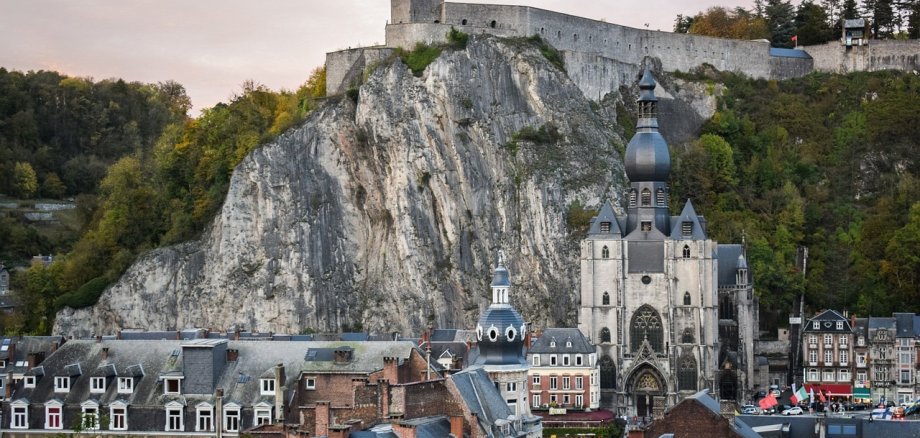 Die Stadt Dinant in Belgien mit ihrer markanten Kollegiatkirche Notre-Dame und den malerischen Häusern im Vordergrund. Darüber erhebt sich eine steile Feldformation mit der Zitadelle von Dinant, die die Szenerie dominiert.
