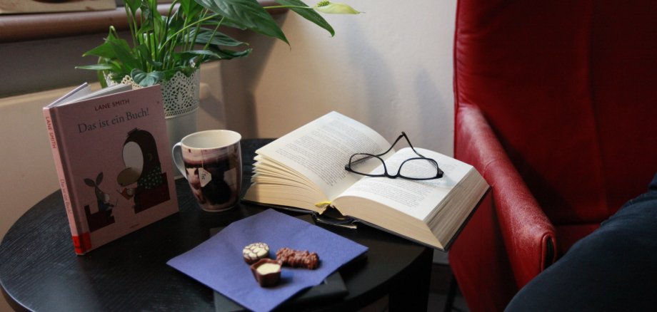 Ein Beistelltisch mit einer Pflanze und einem Bild. Ein großes Buch mit einer Brille liegt auch auf dem Tisch.