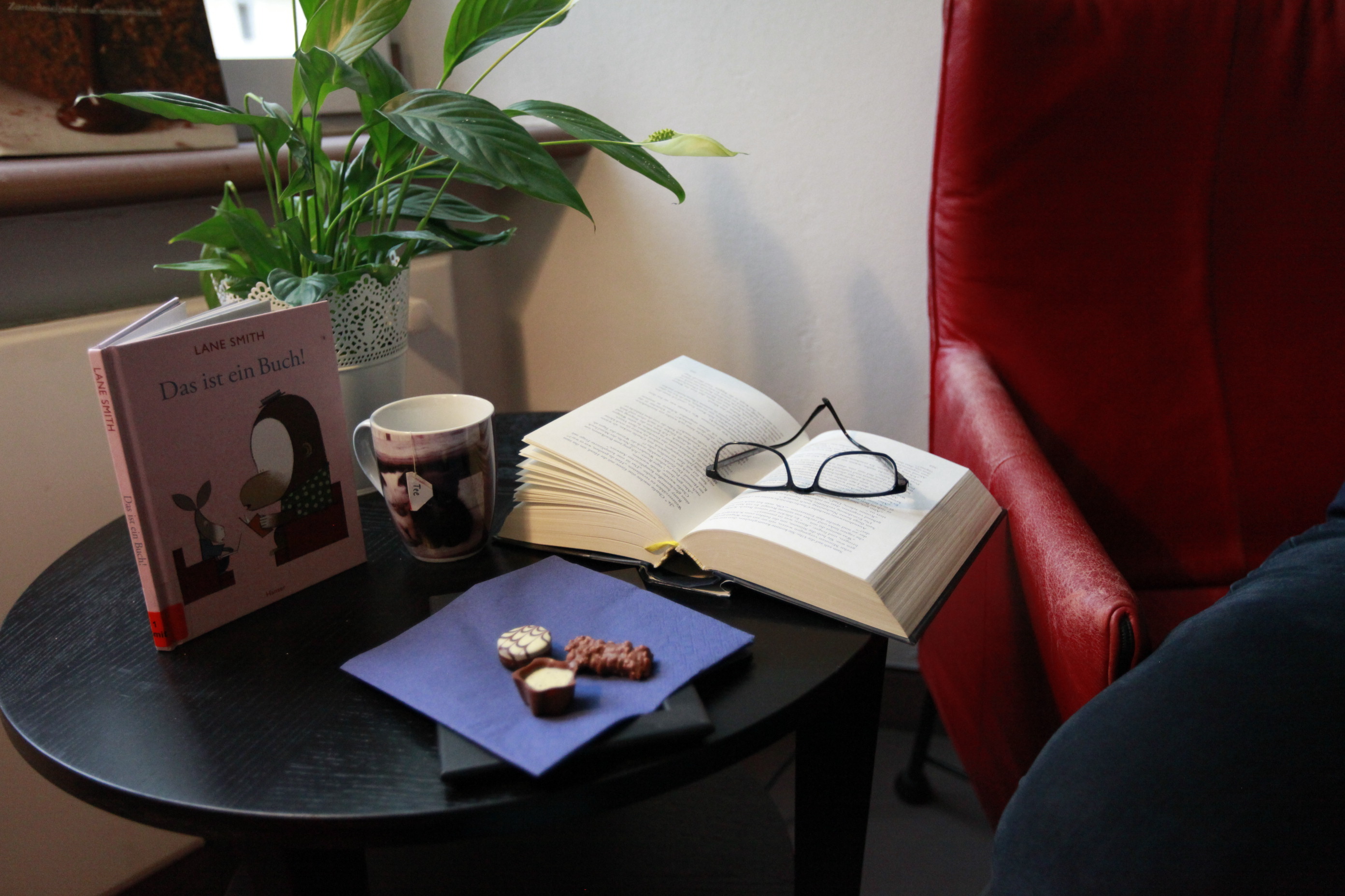 Ein Beistelltisch mit einer Pflanze und einem Bild. Ein großes Buch mit einer Brille liegt auch auf dem Tisch.