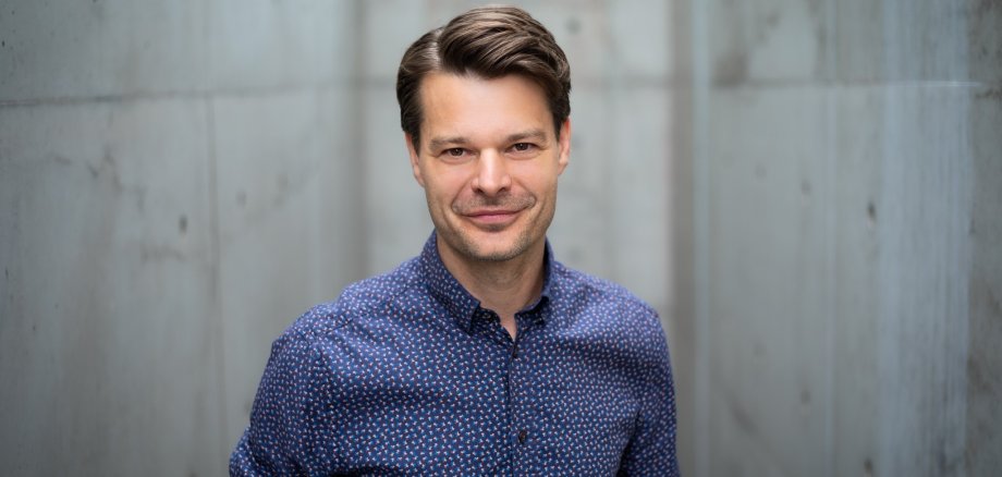 Auf diesem Bild sieht man einen Mann, er ist gut gekleidet mit einem Hemd und gemachten Haaren. Im Hintergrund ist eine Betonwand.
