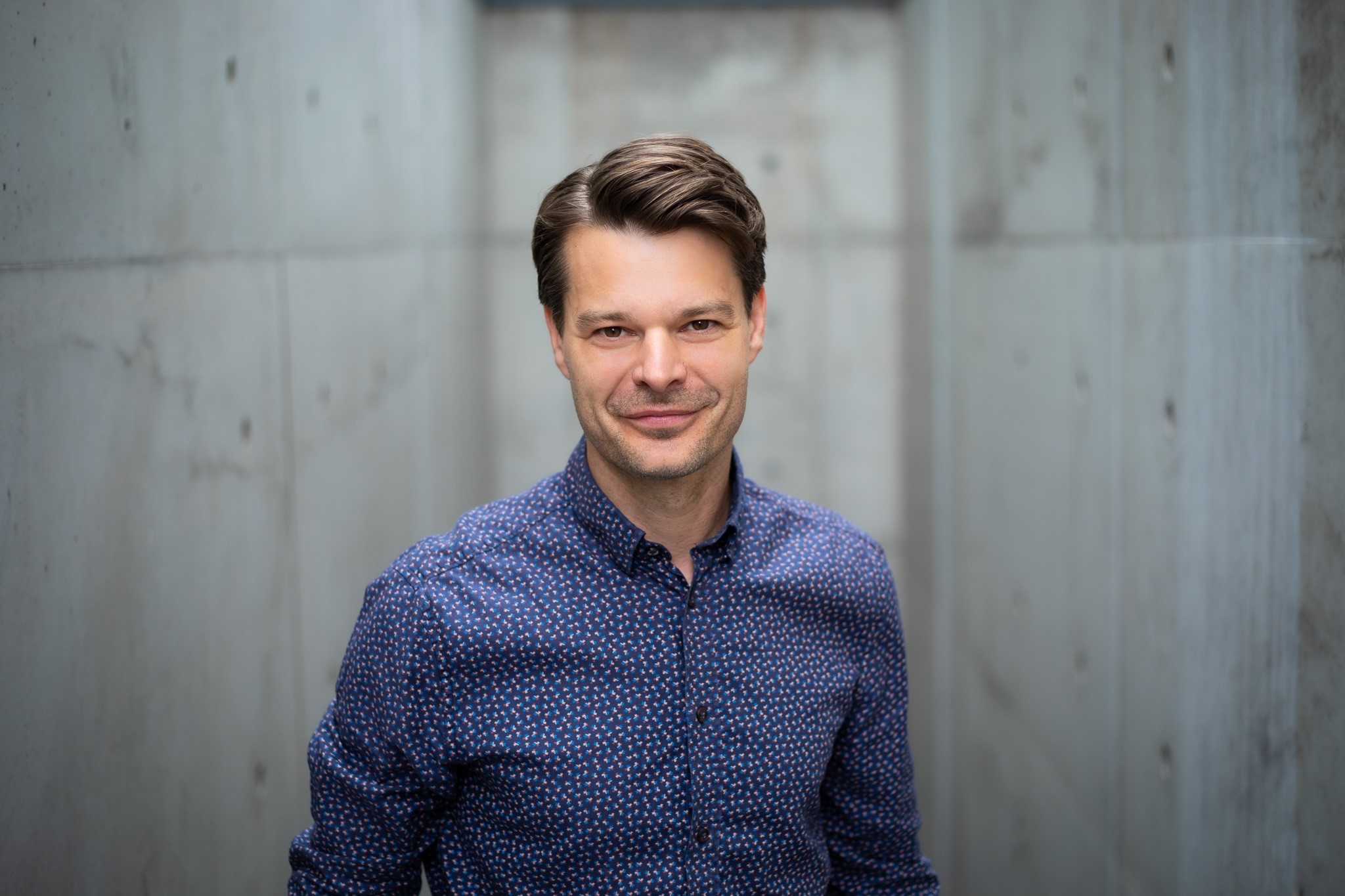 Auf diesem Bild sieht man einen Mann, er ist gut gekleidet mit einem Hemd und gemachten Haaren. Im Hintergrund ist eine Betonwand.