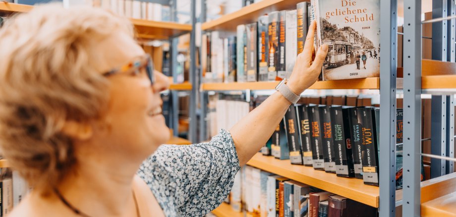 Eine glücklich, lächelnde Frau, welche ein Buch aus dem Regal 1 in der Usinger Stadtbücherei in der Hand hält.