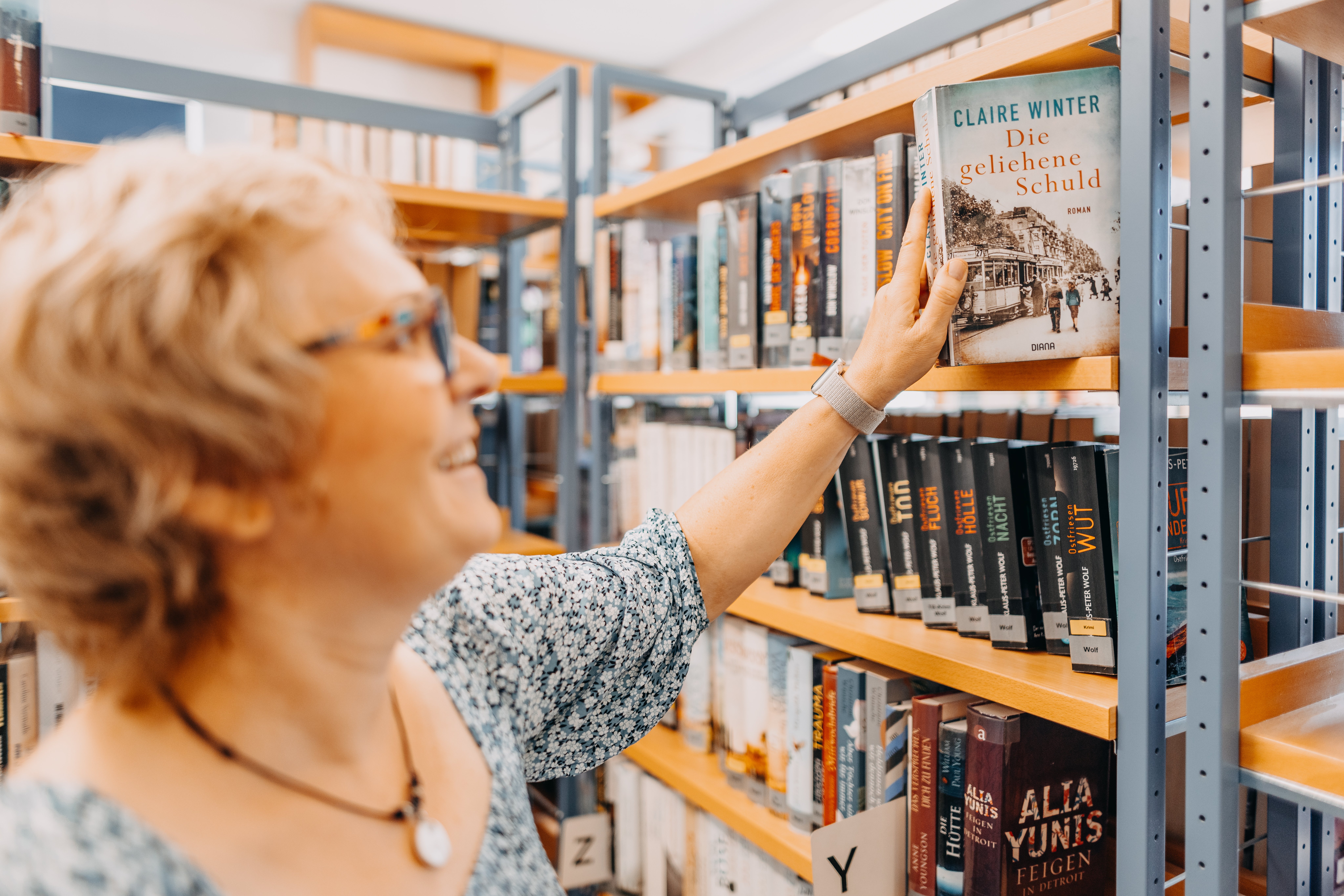 Eine glücklich, lächelnde Frau, welche ein Buch aus dem Regal 1 in der Usinger Stadtbücherei in der Hand hält.