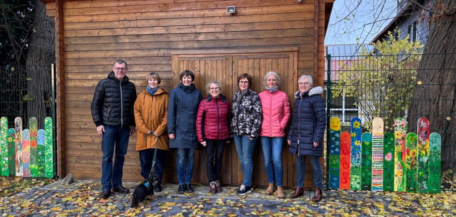 Das Betreuungsteam steht vor einer Holzhütte, links und rechts davon befinden sich die bunt bemalten Zaunlatten.