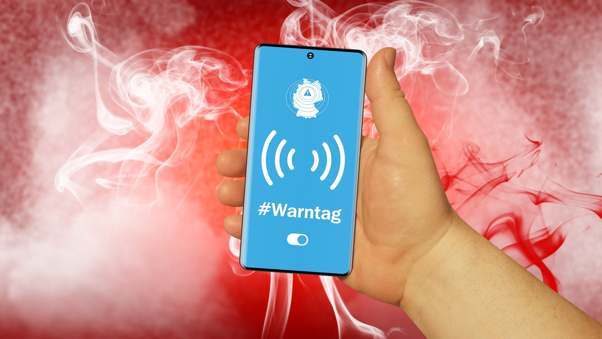 Eine Hand hält ein Smartphone mit blauem Display, auf dem ein Warnsymbol, Funkwellen und der Schriftzug „#Warntag“ zu sehen sind. Im Hintergrund steigen weiße und rote Rauchschwaden auf.