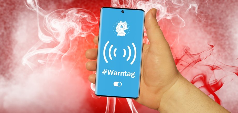 Eine Hand hält ein Smartphone mit blauem Display, auf dem ein Warnsymbol, Funkwellen und der Schriftzug „#Warntag“ zu sehen sind. Im Hintergrund steigen weiße und rote Rauchschwaden auf.
