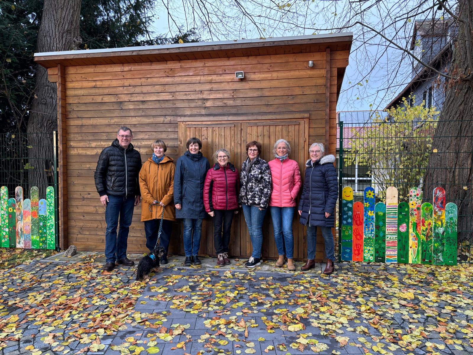 Das Betreuungsteam freut sich über die bunt bemalten Zaunlatten. Das Betreuungsteam steht vor einer Holzhütte, links und rechts davon befinden sich die bunt bemalten Zaunlatten.