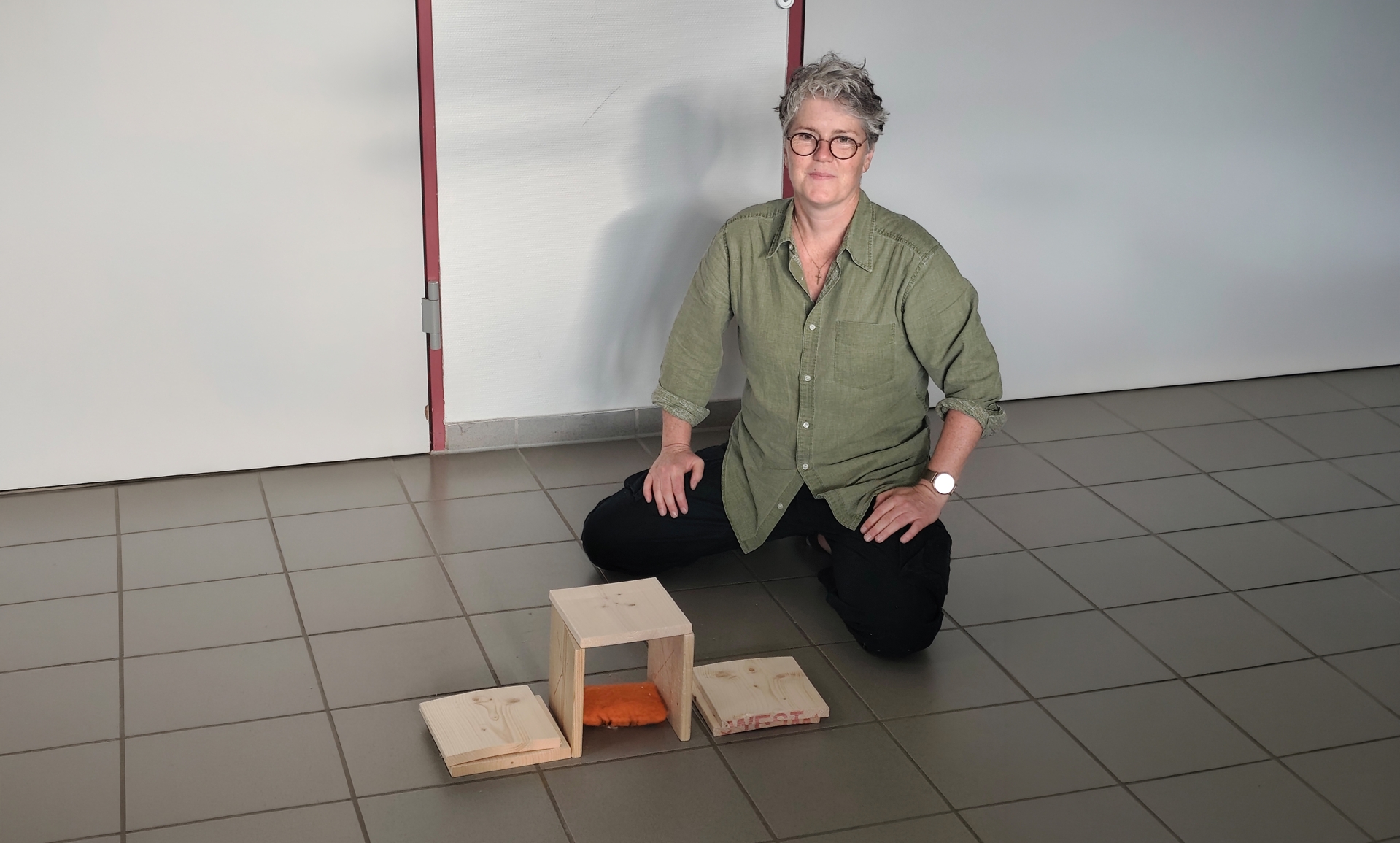 Eine Frau kniet neben einem kleinen selbst gebauten Holzobjekt auf dem Boden und blickt in die Kamera.