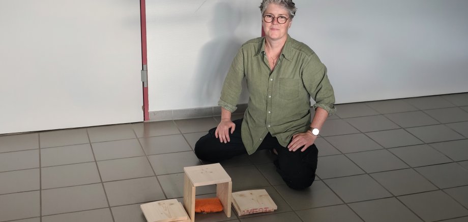 Eine Frau kniet neben einem kleinen selbst gebauten Holzobjekt auf dem Boden und blickt in die Kamera.