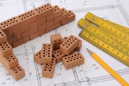 Bauleitplanung Auf einem Bauplan liegen ein gelber Zollstock, ein gelber Bleistift und Backsteine im Miniaturformat.