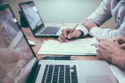 Büro Zwei Menschein sitzen an zwei Laptops. Zwischen den beiden Laptops liegen Unterlagen.